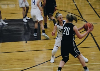 Two female basketball players compete in a basketball game. One is wearing a white uniform, and is guarding a player in a black uniform. 