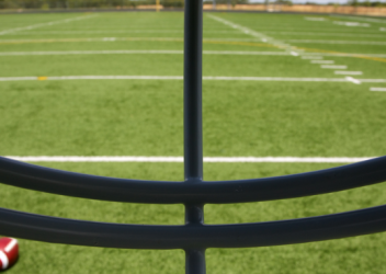 A football field is seen through the vantage point of a player looking through a helmet facemask. A football is on the ground to the left of the player. 