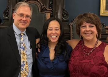 A photo of a room with a dark blue wall and a long table in the background with dark brown chairs that are tall. In the forefront is Dr. Matthew Grady in a dark suit, light blue shirt, and yellow, orange and blue tie; Dr. Christina Master, wearing a blue dress, and Dr. Kristy Arbogast, wearing a red dress. 