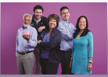 Five members of the Minds Matter Concussion Program are seen smiling straight ahead at the camera, in front of a purple-colored backdrop. The person on the left is wearing a light-blue dress shirt and is holding a pink-colored model of a brain. 